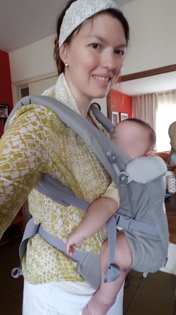 Josie Zecchinelli em frente ao espelho com bebê em mochila ergonômica cinza com apoio postural e cinto lombar