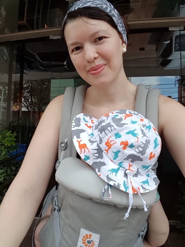 Josie Zecchinelli em passeio ao ar livre com bebê de chapéu estampado na mochila ergonômica cinza